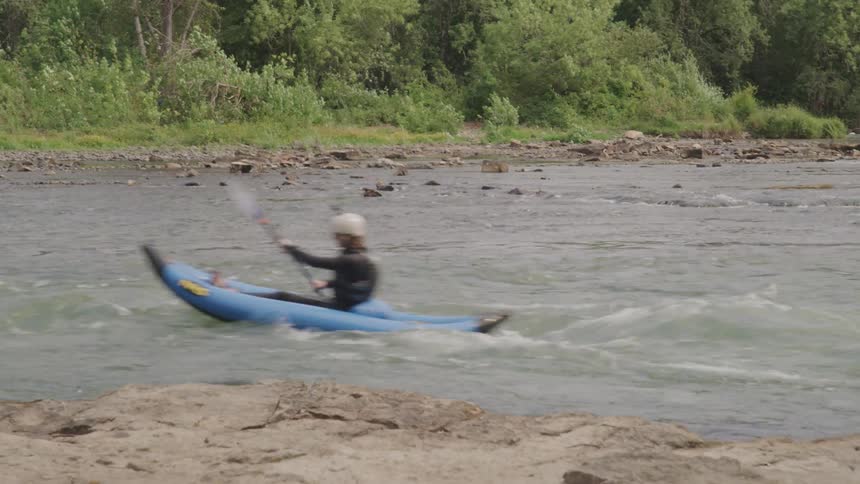 A person in a blue kayak is navigating through mild rapids of a river with a forest in the background