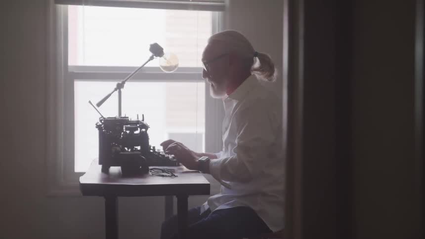 Older man types on mechanical typewriter near window