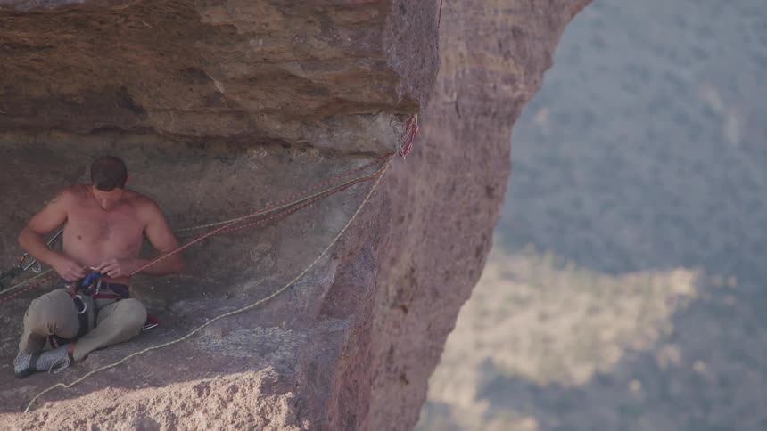 A rock climber rests on a ridge to adjust his ropes