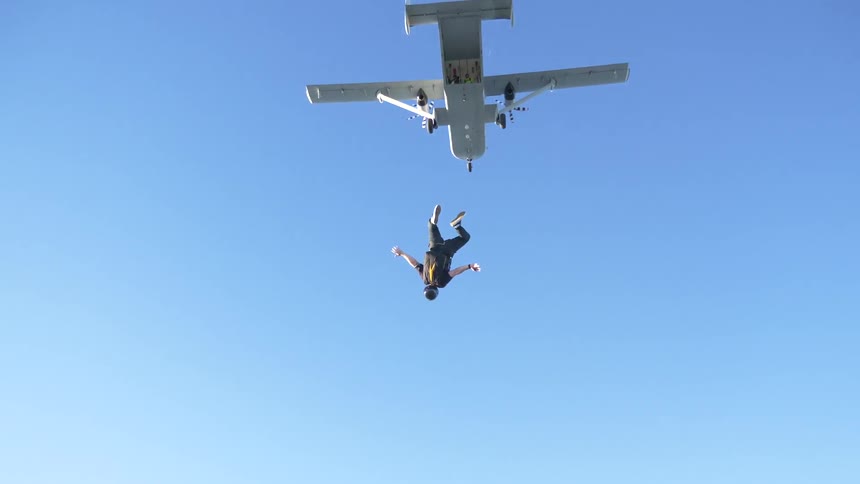 A man with a parachute backpack steps out of the opening of a skydiving plane tucks his knees and does two somersaults before opening his arms and legs to fall