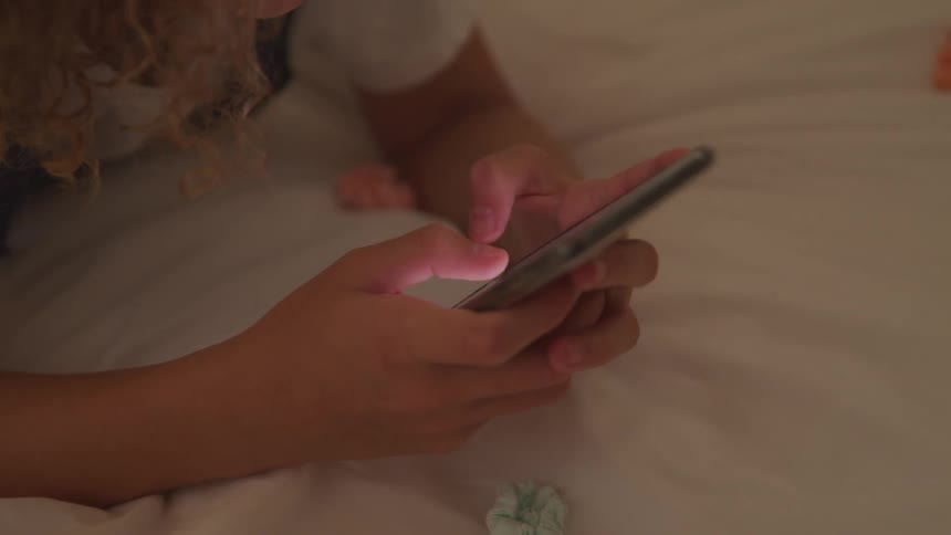 Girl lying down on the bed uses the smartphone in the room