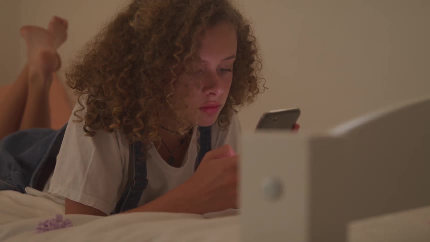 A girl using her smartphone while resting on a bed
