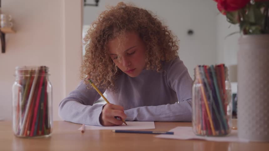 Curly red hair girl sitting at the table draws on paper with pencil in a room