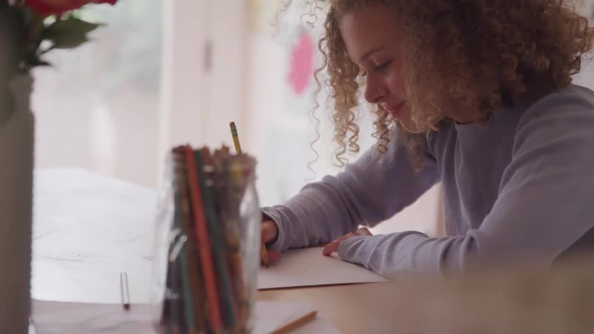 Red curly hair girl sitting at the table draws on the paper in a room