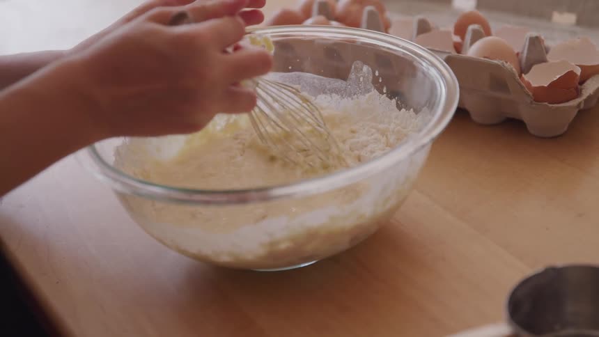 A person is mixing ingredients in a glass bowl using a metal whisk with a carton of eggs off to the side on the counter