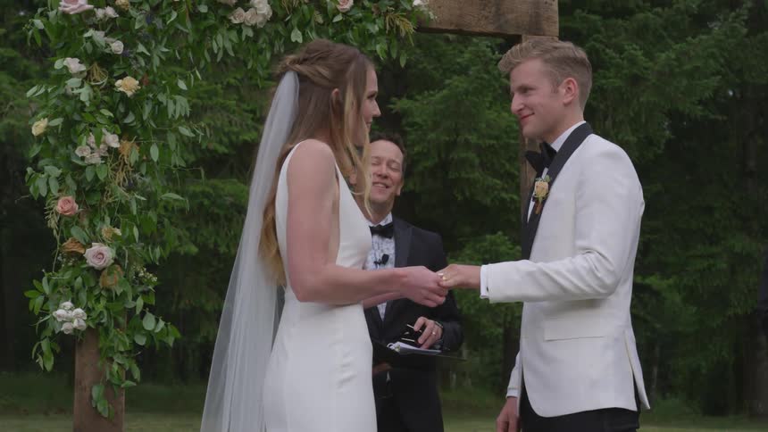 A bride and groom hold hands and smile at each other as a man standing behind them speaks