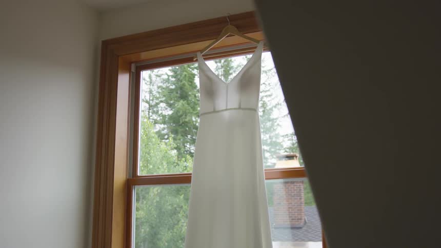 A wedding gown hangs front a hanger in front of a window