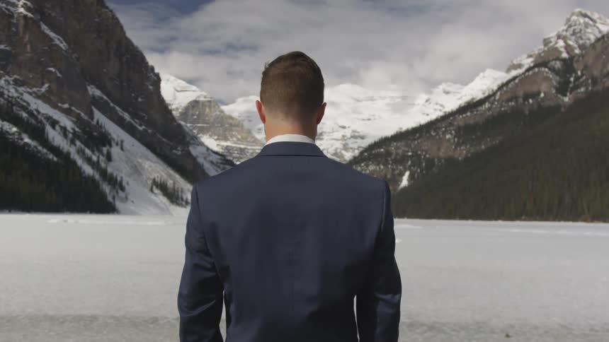 A man in a suit coat stands facing away towards a winter landscape set in a valley between towering snow covered mountains