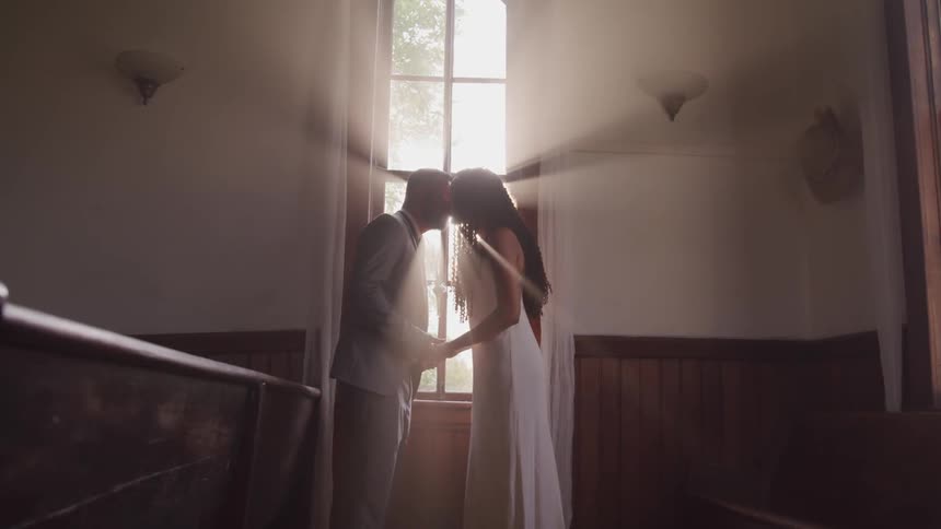 Bride and groom kissing by a sunlit window in a church