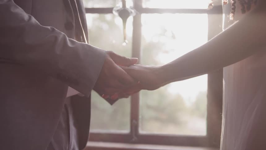 Groom and bride hold hands and look at each other while the sun streams through the window during wedding ceremony