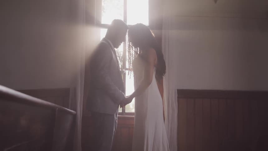 Groom and bride hold hands and look at each other while the sun streams through the window