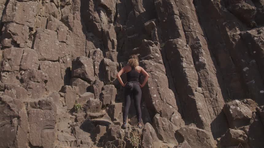 A woman stands on a rocky cliff looking at the mountain in front of her