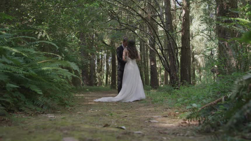 A bride and a groom stand facing each on trail in the middle of a forest