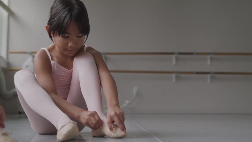 Two little ballerinas sitting on the ground put on shoes in the ballet studio