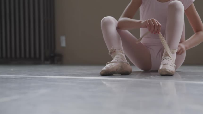 A young ballerina sits on the floor wrapping the ribbons of her ballet shoes around her leg to tie