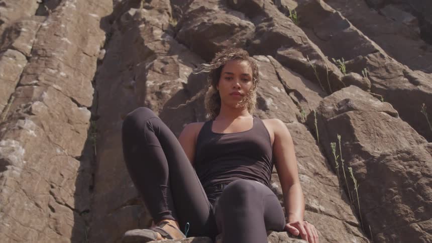 A woman wearing fitness apparel sits atop a rock at the base of a towering rocky cliff