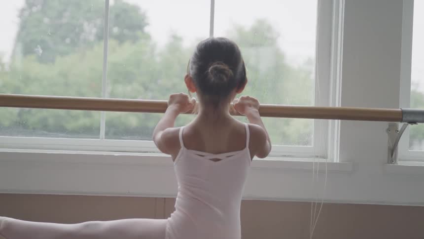 Young ballerina holds onto a barre while looking out the window and lifts her left leg out to the side and then out behind her