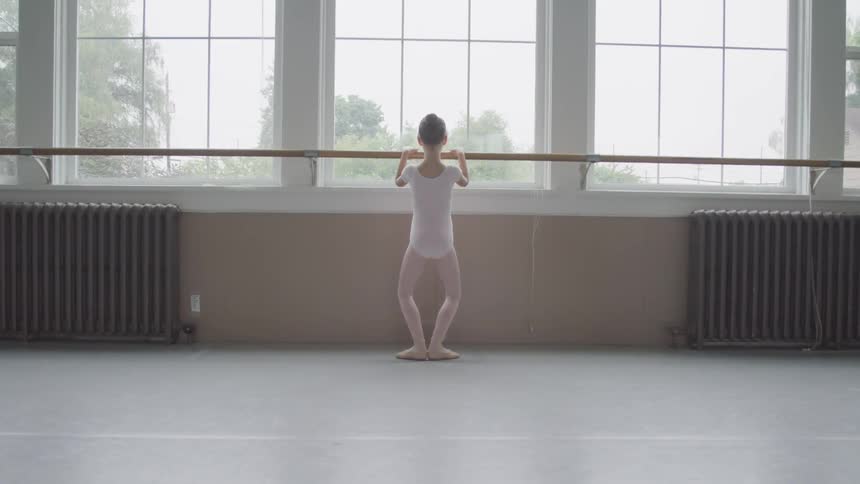 A young ballerina is practicing ballet with her feet in first position in front of a barre in a studio