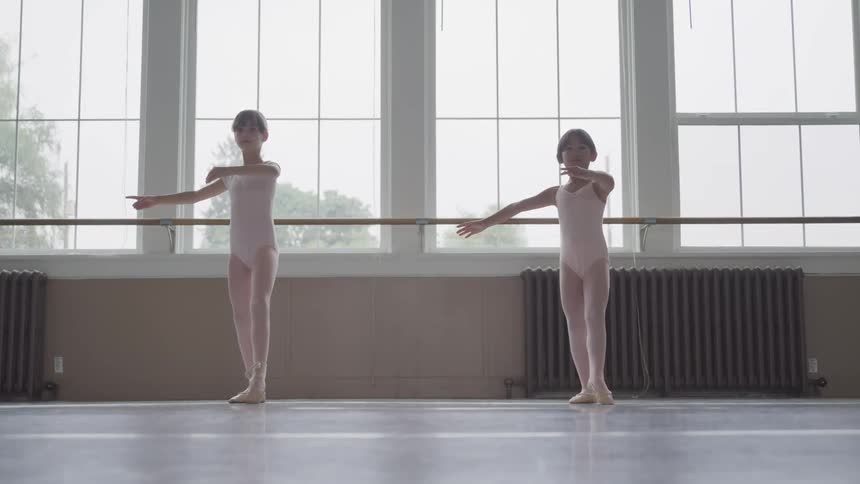 Two young girls in ballet slippers and leotards are practicing dance moves together in a studio with a wall of windows