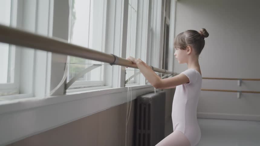 Female child ballerina has both hands on a barre in a dance studio while looking out the window and lifts her leg into a figure four position