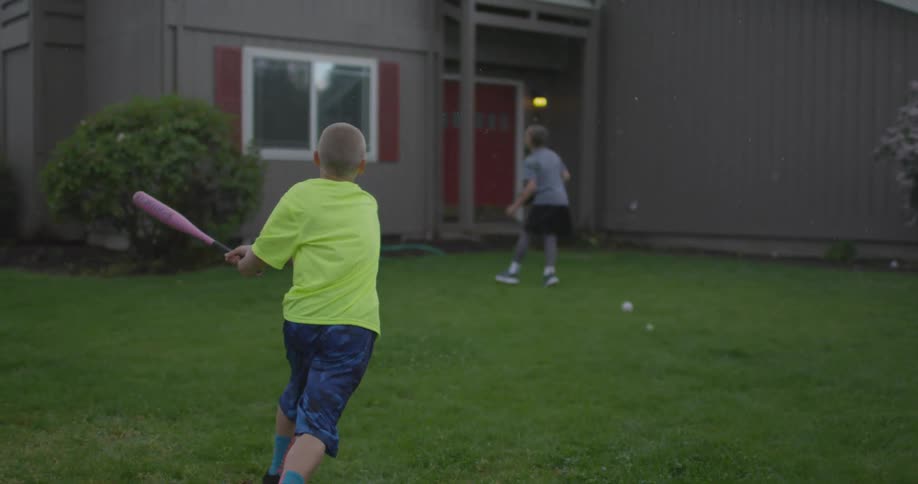 Two boys play baseball in the front yard of a home and the struck ball almost hits a window