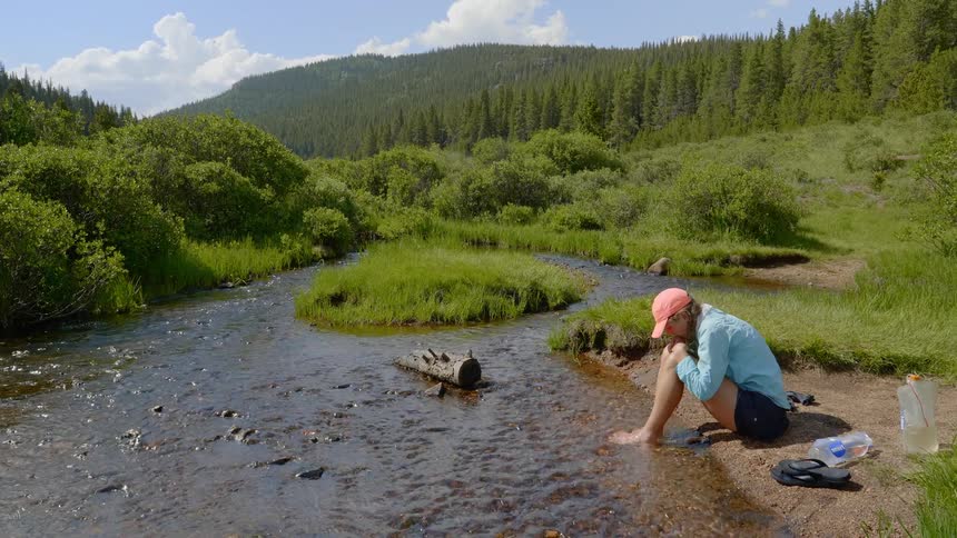 A white female is sitting on the ground with her feet in a stream with evergreens and mountains in the background