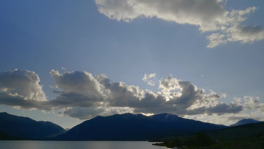A mountain range with fluffy clouds above it obscuring the sun at the edge of a lake