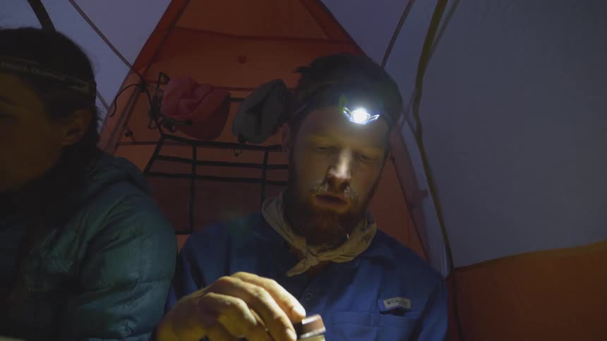 A man sitting in a tent opens a tin and looks at the contents inside