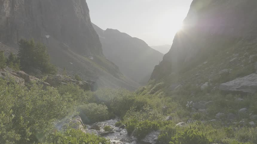 A stream runs through a valley lit by the sun thats being partially blocked by a mountain