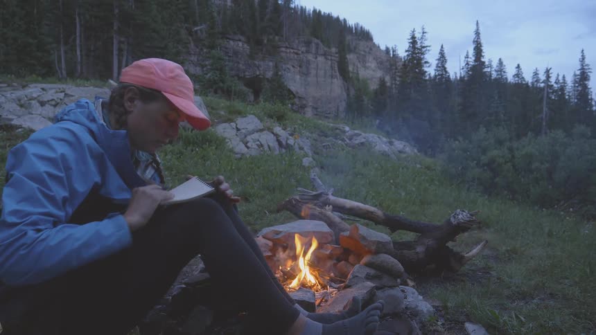 Woman sitting on the ground next to the campfire writes down in the forest at dusk