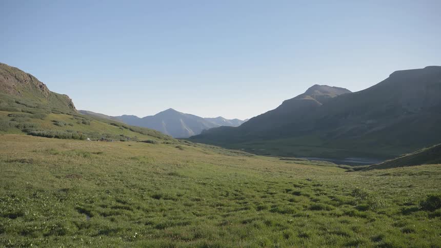 A large grass filled valley sits below a rocky mountain range