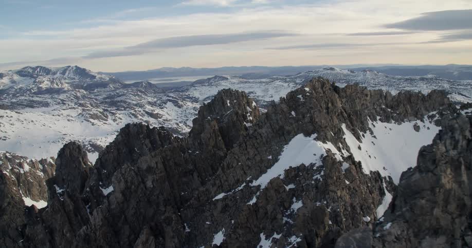 Aerial footage of jagged, snow-capped mountains peaks under a cloudy sky