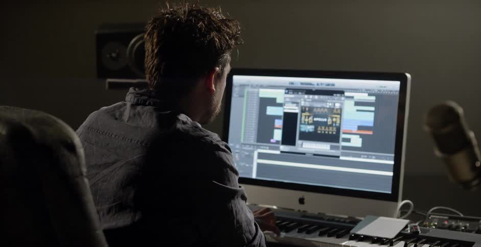 A white male is sitting at a desk using a keyboard and apple computer to make music