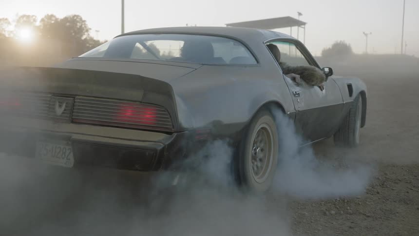 Two people in a car as the back wheel turns causing dust to raise and the car to smoke