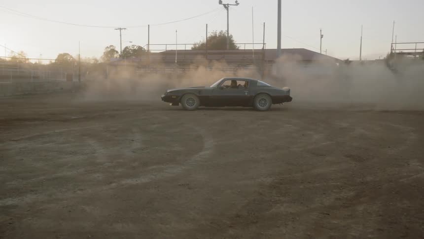 A classic sports car accelerates and then takes a sharp turn and spins around in a dusty outdoor field next to some bleachers