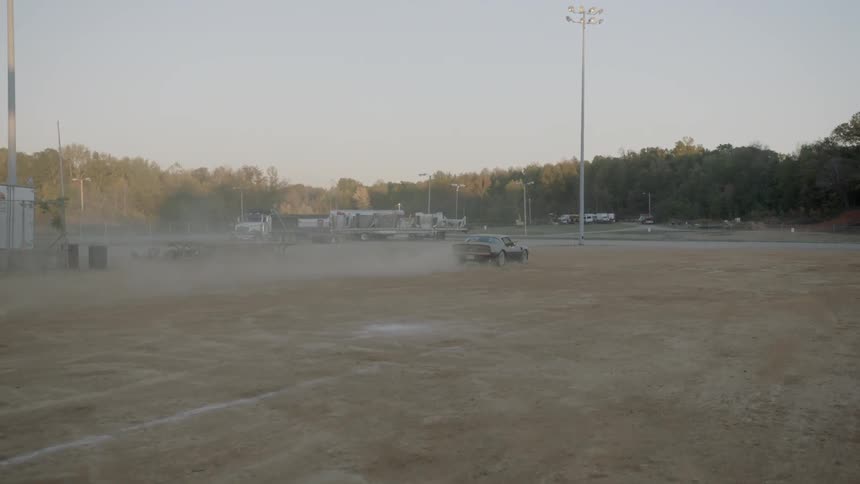 A classic sports car drives through a wide open dirt field while kicking up a cloud of dust behind itself