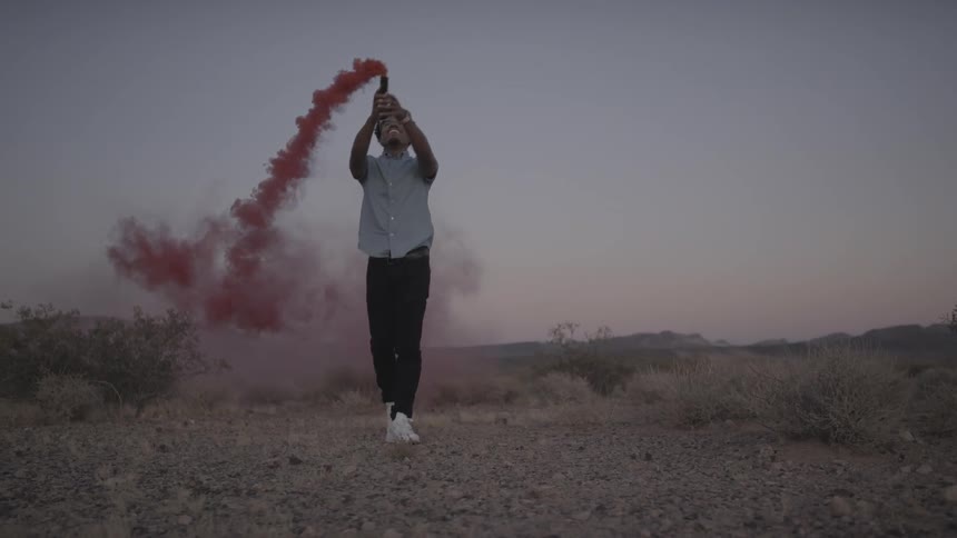 Man in light shirt and dark pants waves device emitting red smoke outside in a field