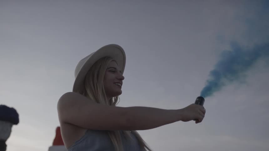 A young woman in a hat stands outside and smiles as shes releasing blue smoke from a small canister
