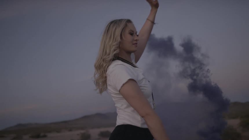 A blonde woman spins around while holding a smoke bomb