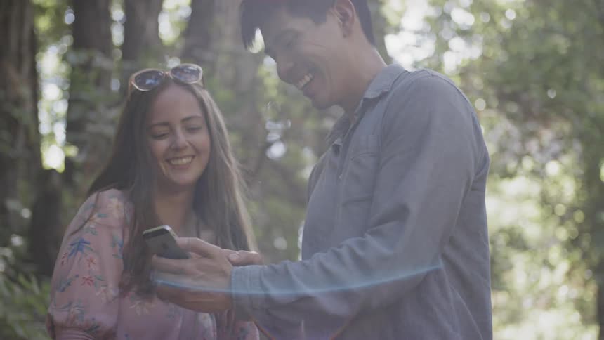 A man and a woman laugh as they stand in a forest while looking at a cell phone