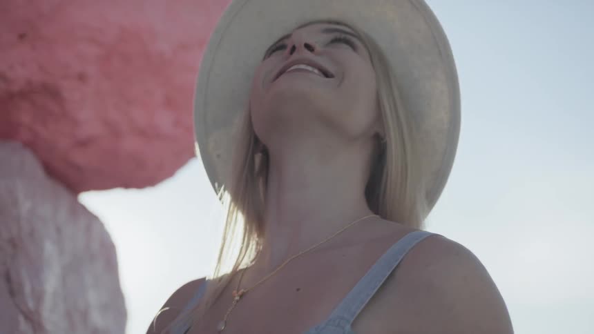 A young blonde woman wearing a hat stands outside next to some large rocks and smiles while looking upwards