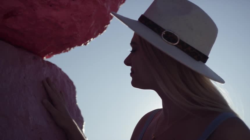 Blonde woman wearing wide hat touches colorful stone pillar in sunny weather