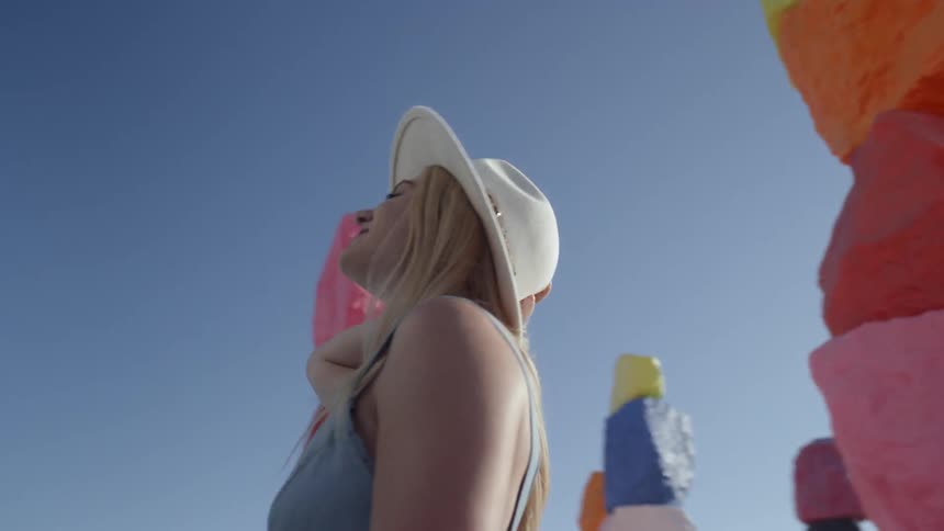 A woman holds her hat on her head as she looks up at brightly painted stacked rocks in the desert