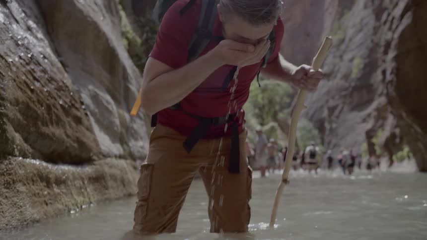 A hiker walks through water in a canyon and drinks from his cupped hands