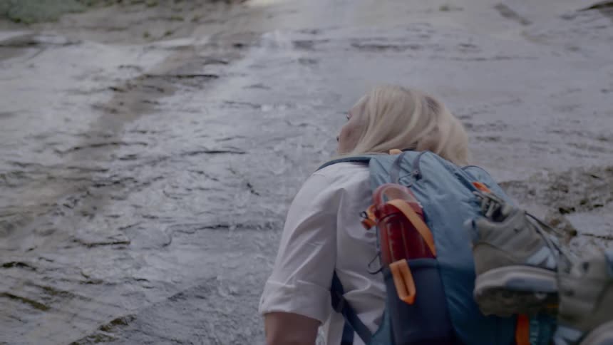 A woman with blonde hair wears and blue backpack and bends over over to observe water running along the ground