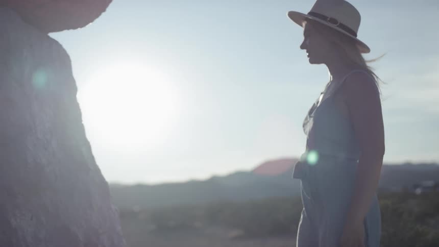 A woman in a sun hat walks up to a large boulder and touches it