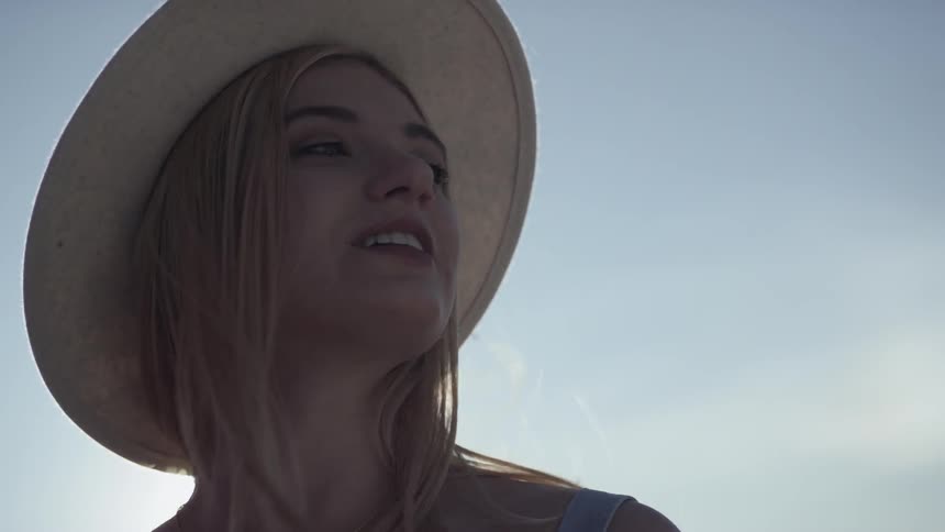 Blonde woman wearing wide hat looks around while the sun shines behind