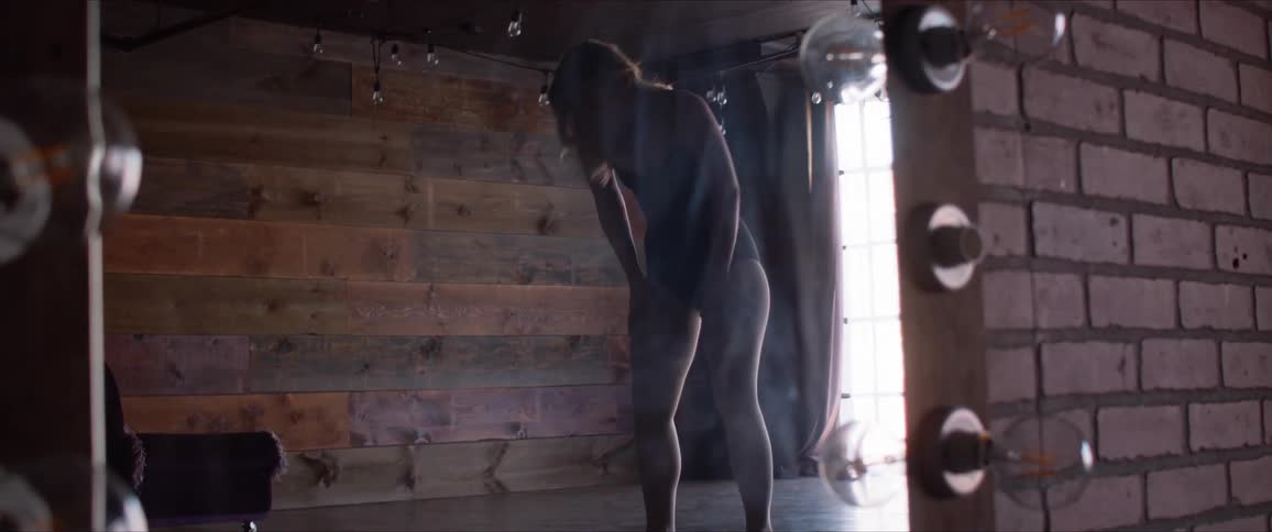 A woman in a ballet attire stands before a mirror and leans over to catch her breath
