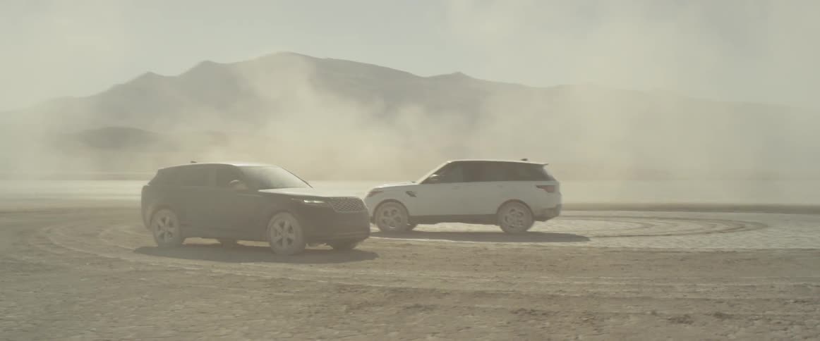 Two sport utility vehicles are parked atop a dried up and dusty lake bed as clouds of dust linger around them