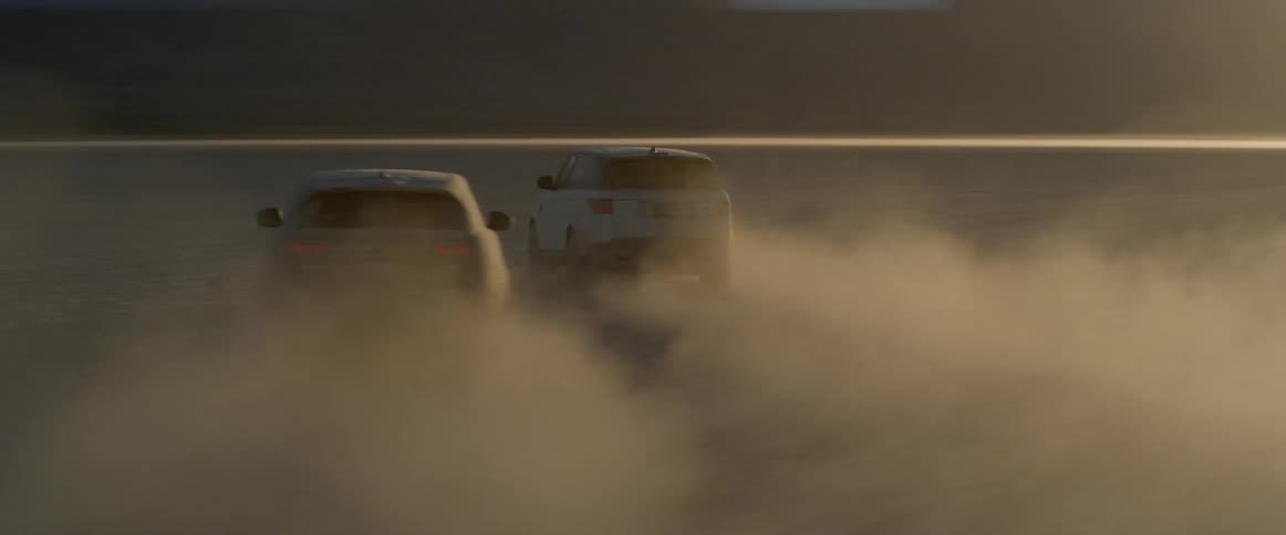 A black car and white car are driving through some desert flats at sunset